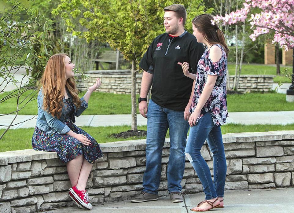 Three people, two standing and one sitting on a stone ledge, 在盛开的树木和绿色植物附近进行户外对话.